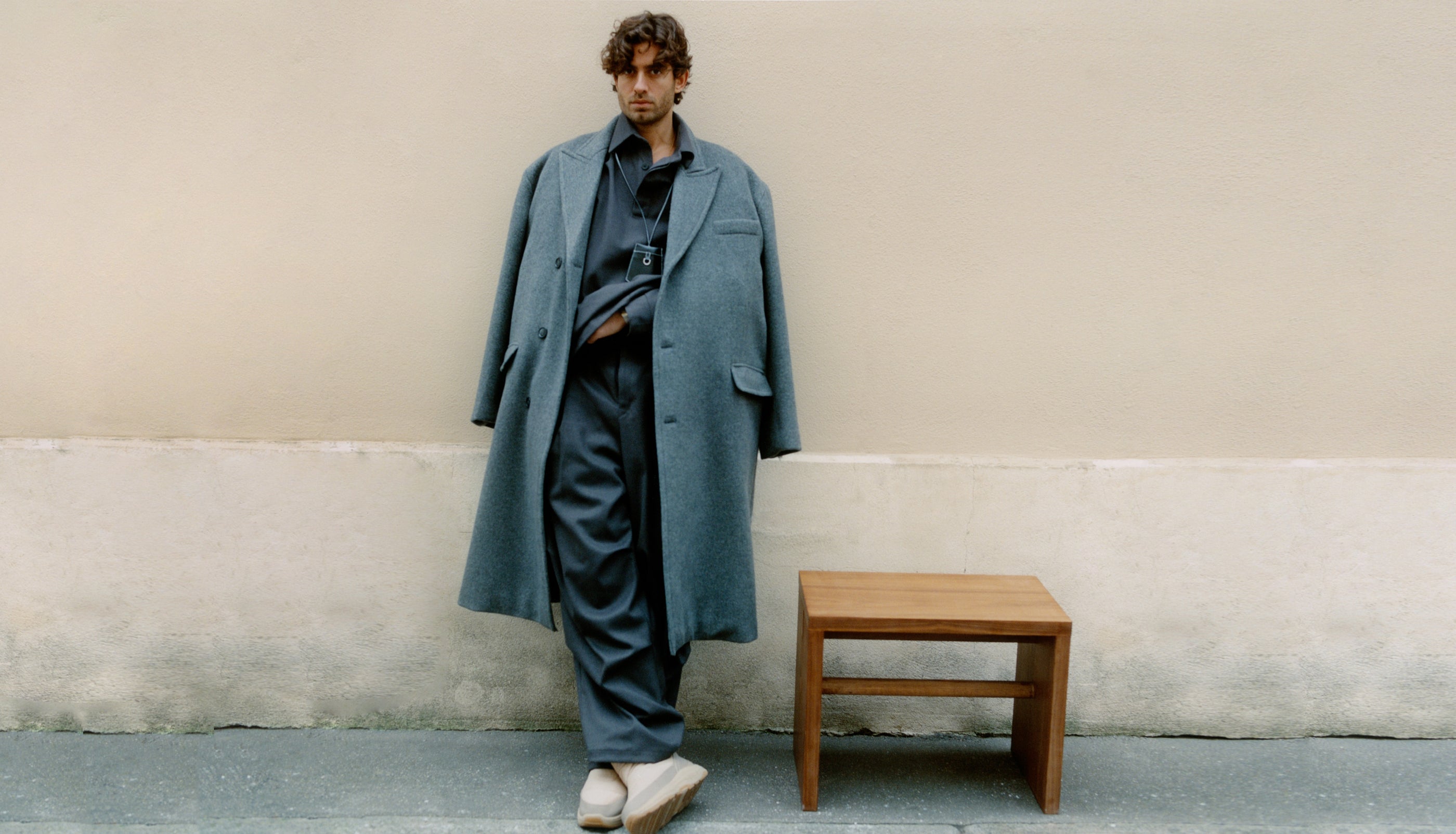 Male model standing against a wall photographed by Deon Hinton wearing the Curtis coat and Flint pants by The Frankie Shop. 