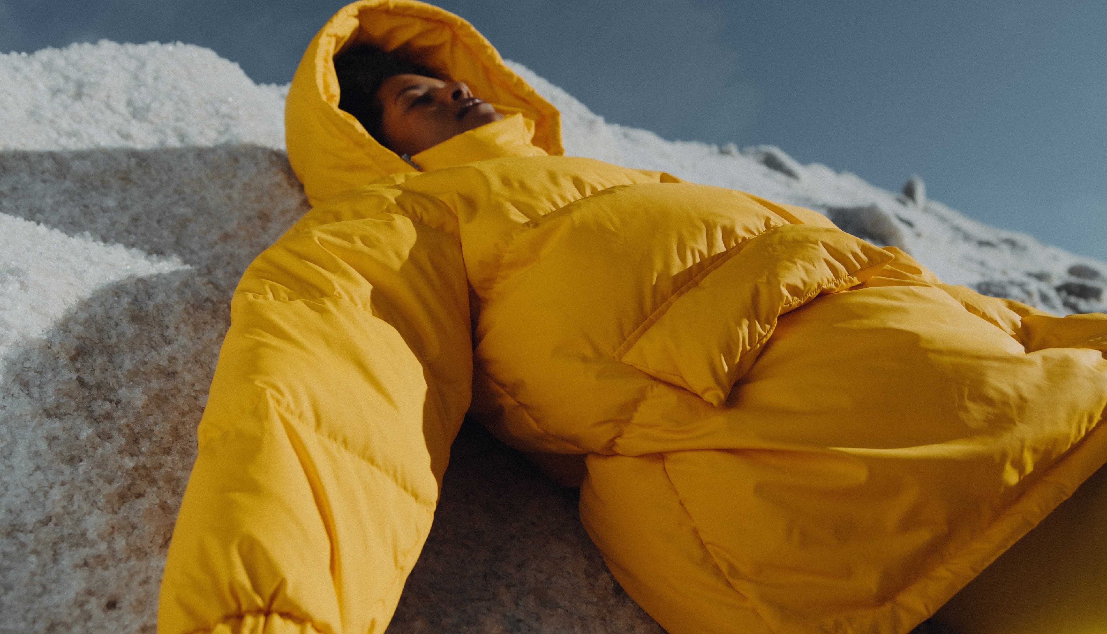 Model lying on the snow capped mountain wearing the Val puffer jacket in yellow. Photographed by Charlotte Lapalus
