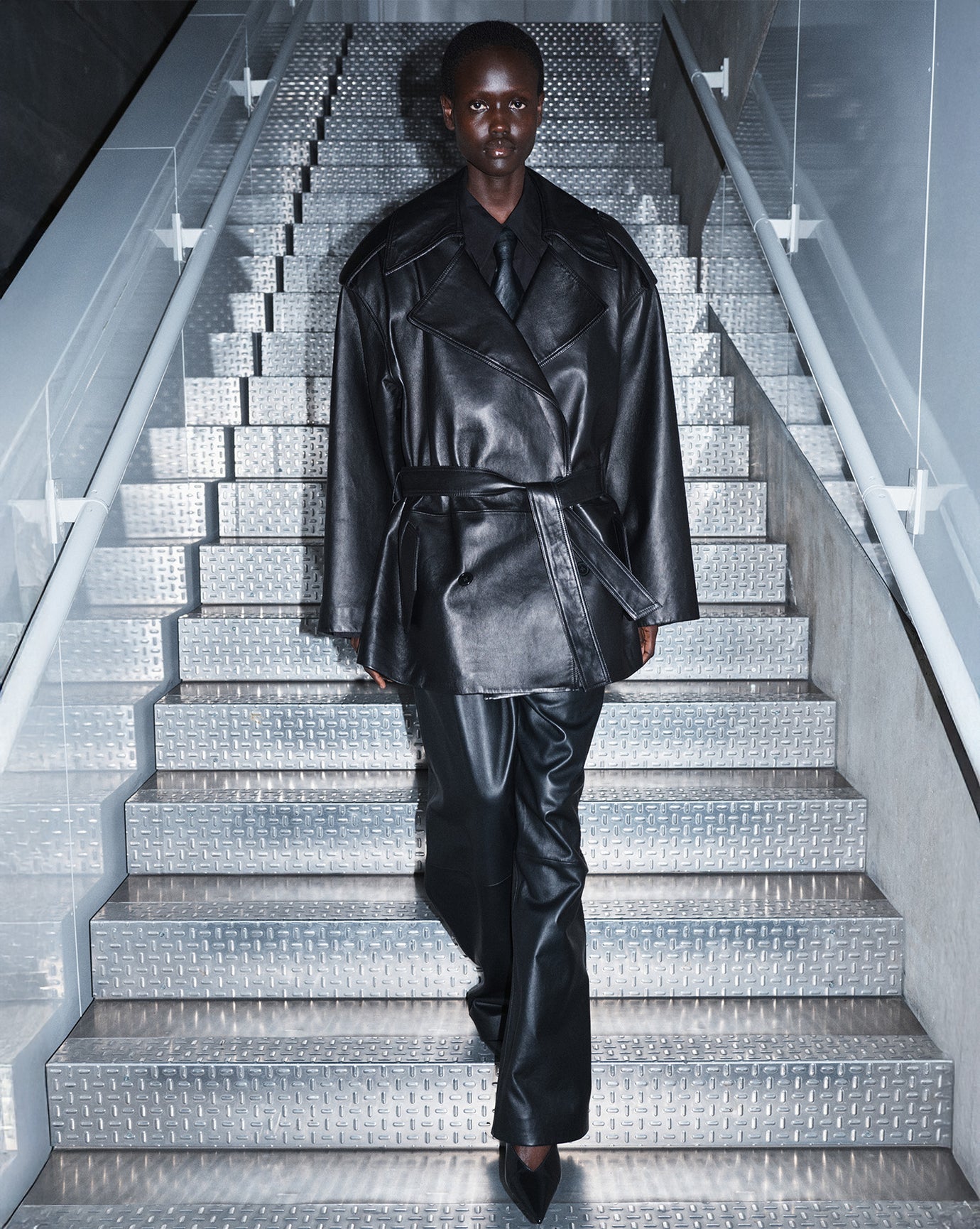 Model photographed against a green backdrop wearing a grey tailored suit holding a large black leather bag.