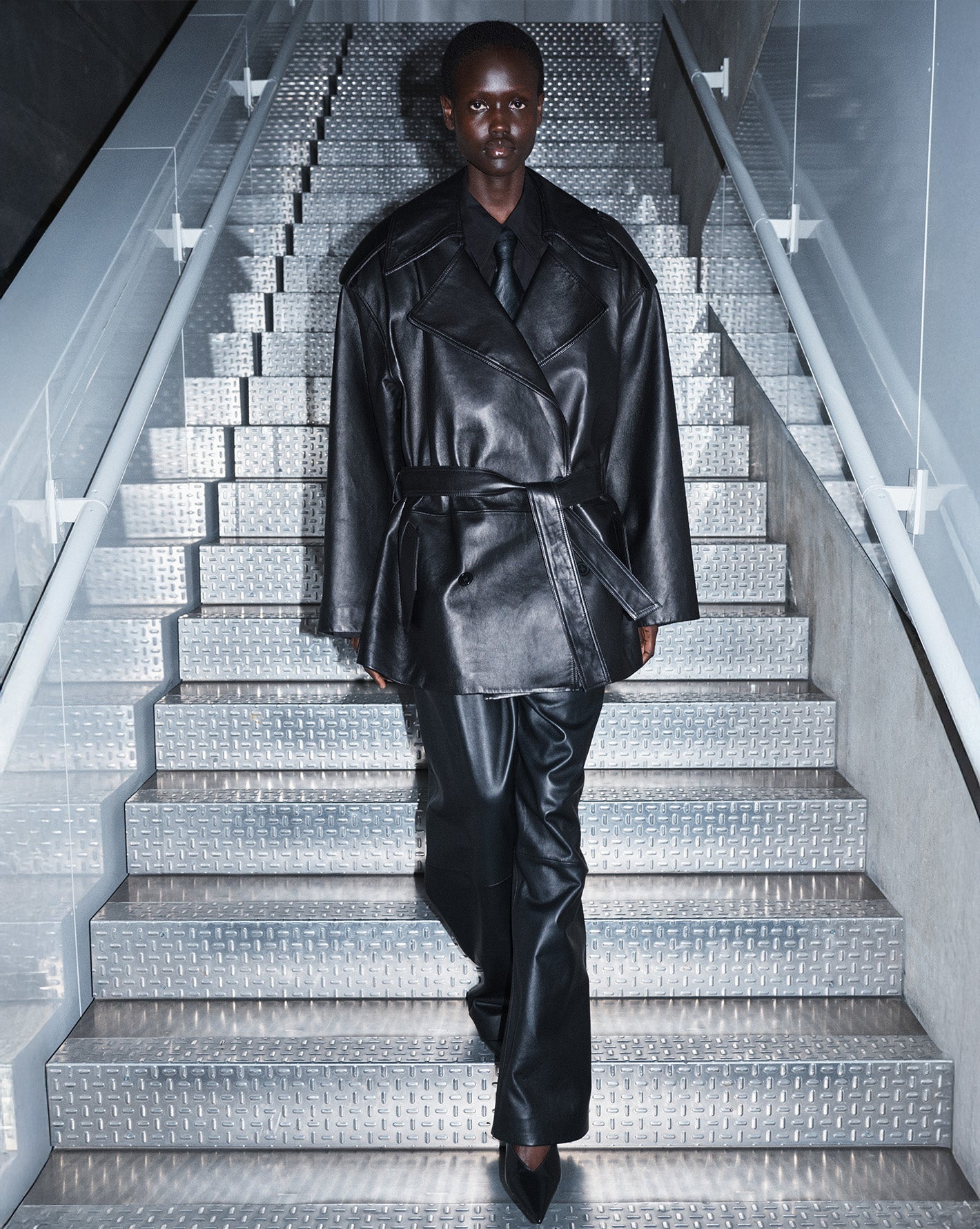 Model photographed against a green backdrop wearing a grey tailored suit holding a large black leather bag.
