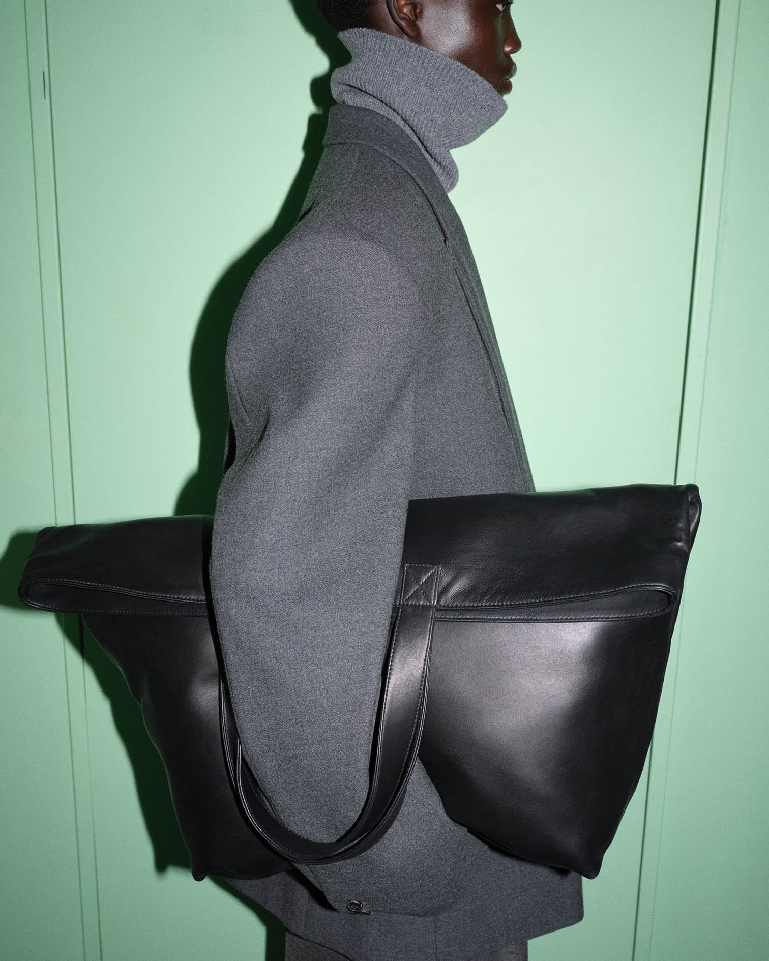 Model photographed against a green backdrop wearing a grey tailored suit holding a large black leather bag.